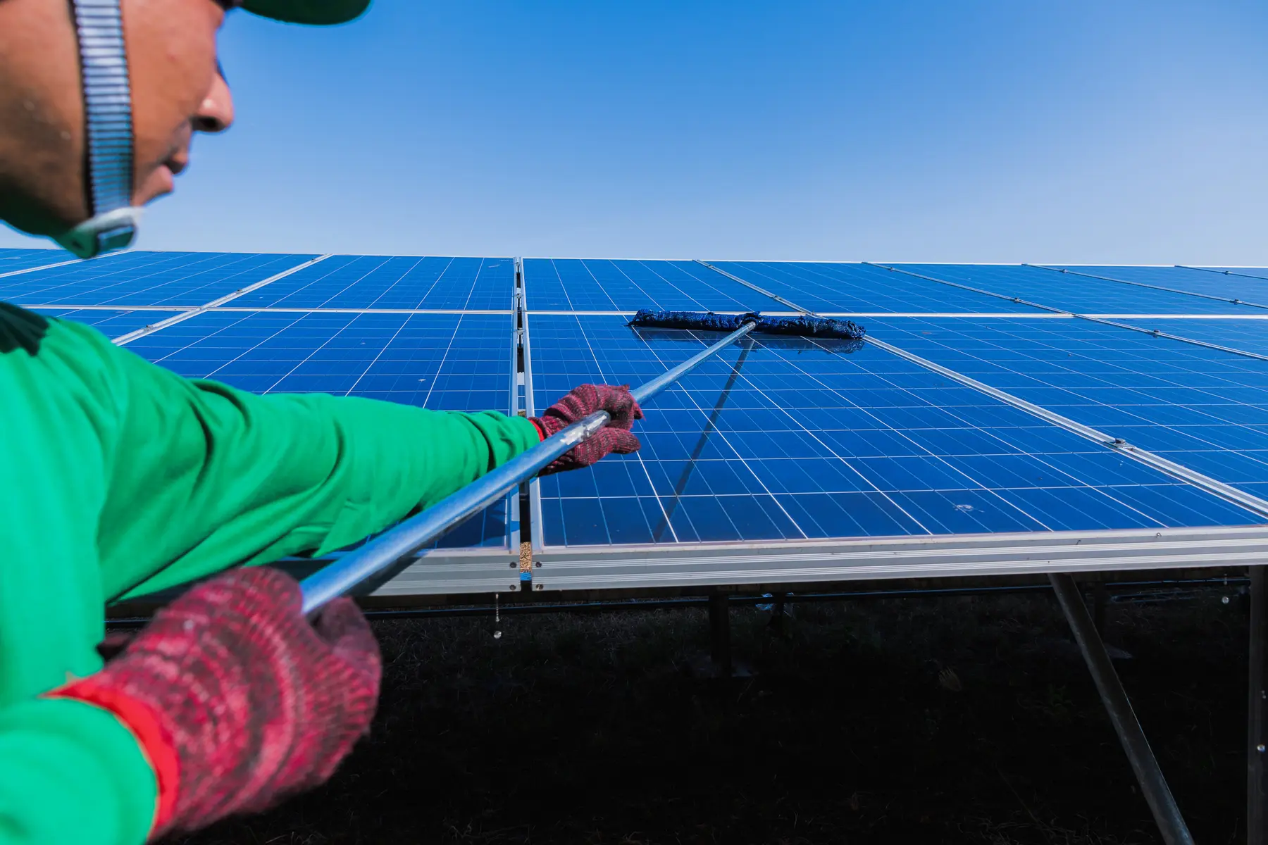 Operario limpiando con pértiga y cepillo una hilera de placas solares, con el agua escurriendo por el vidrio